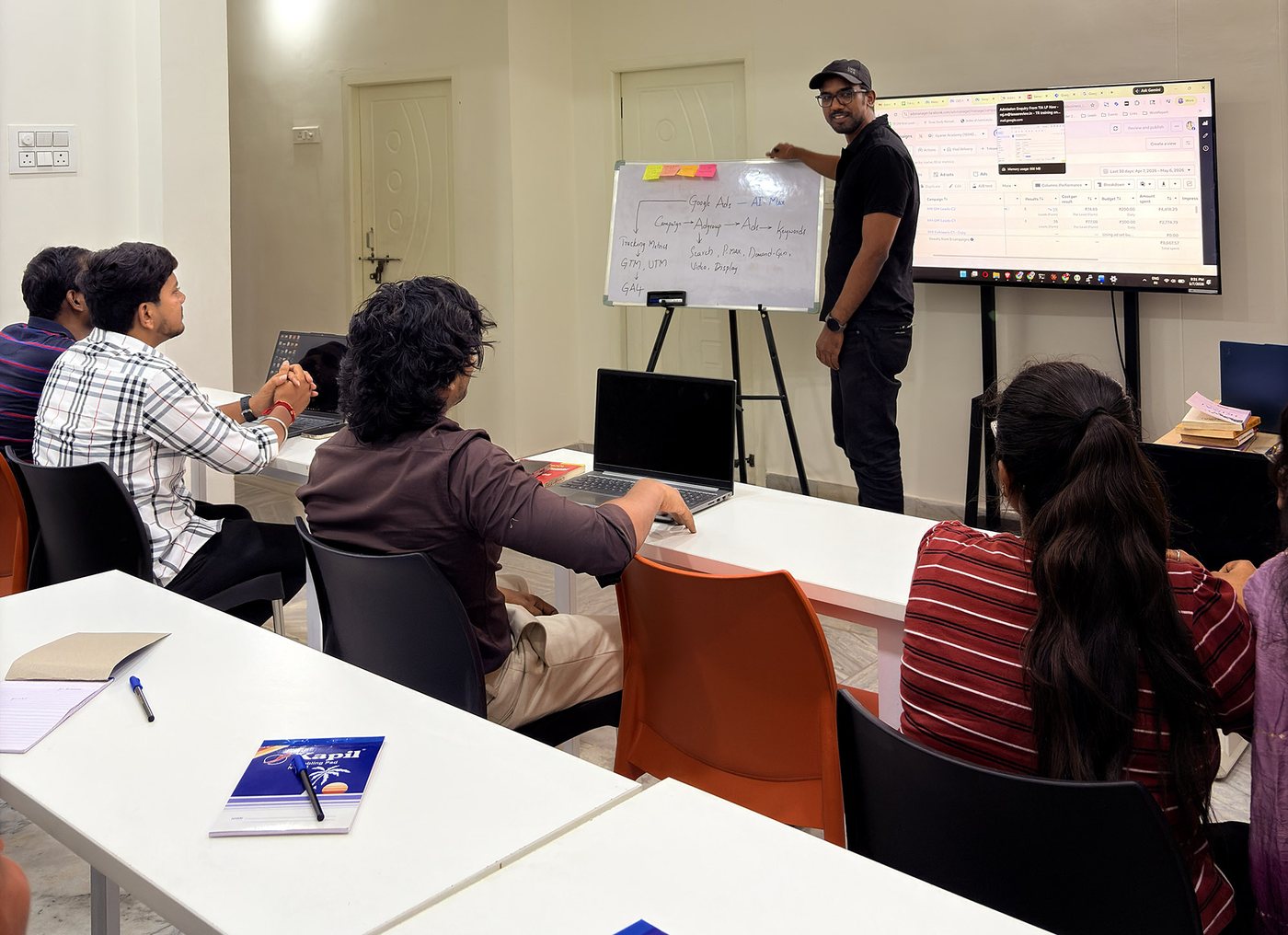 Trainer explaining Google Ads AI Max campaign structure on a whiteboard beside a live ads dashboard, students working on laptops at Gyaner Academy in Hyderabad